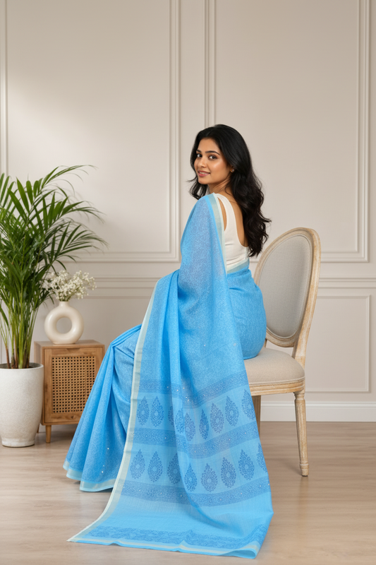 A woman modeling a light blue saree with subtle tonal patterns and a matching blouse, standing in a neutral indoor room with a plant and small wooden side table.