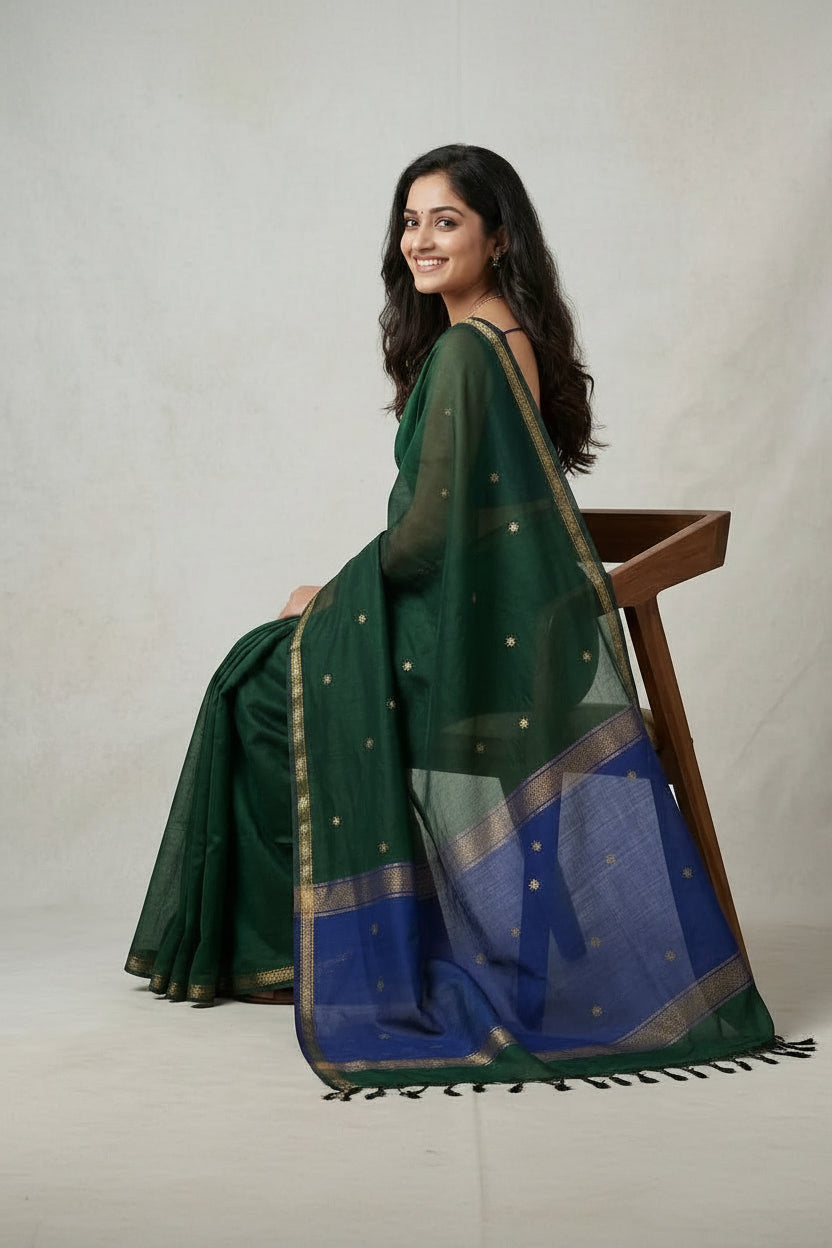 Woman wearing a green and blue saree sitting on a wooden stool against a plain background