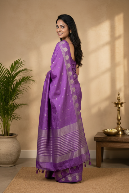 Woman in a purple saree with silver patterns standing in a room with a plant and small table.