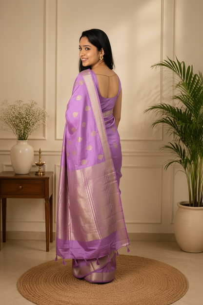 Woman wearing a purple saree with white polka dots in a room with plants and a wooden side table.