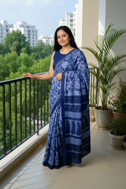 A woman wearing a blue patterned saree and matching sleeveless blouse stands on a balcony with green trees and buildings in the background.