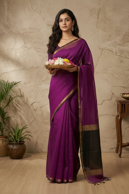 Woman wearing a purple saree with a gold border against a beige stone wall.