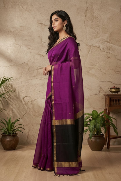 Woman wearing a purple saree with a gold border against a beige stone wall.