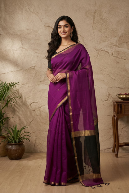 Woman wearing a purple saree with a gold border against a beige stone wall.