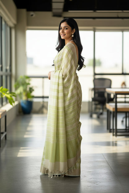 Woman in a light green saree standing in an indoor setting with large windows.