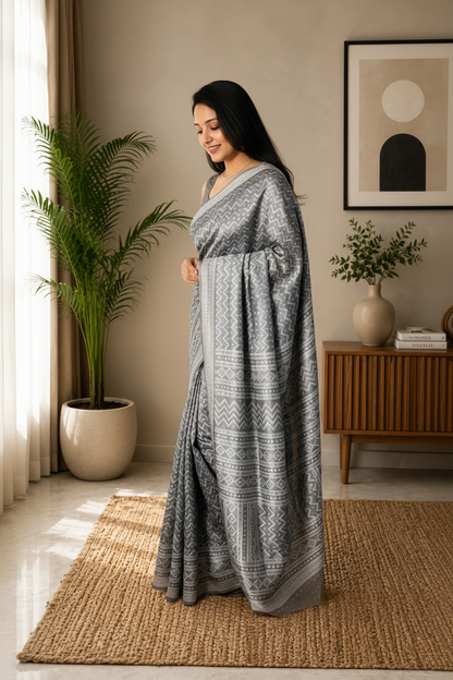 Woman in a grey saree standing in a room with plants and a wooden cabinet.