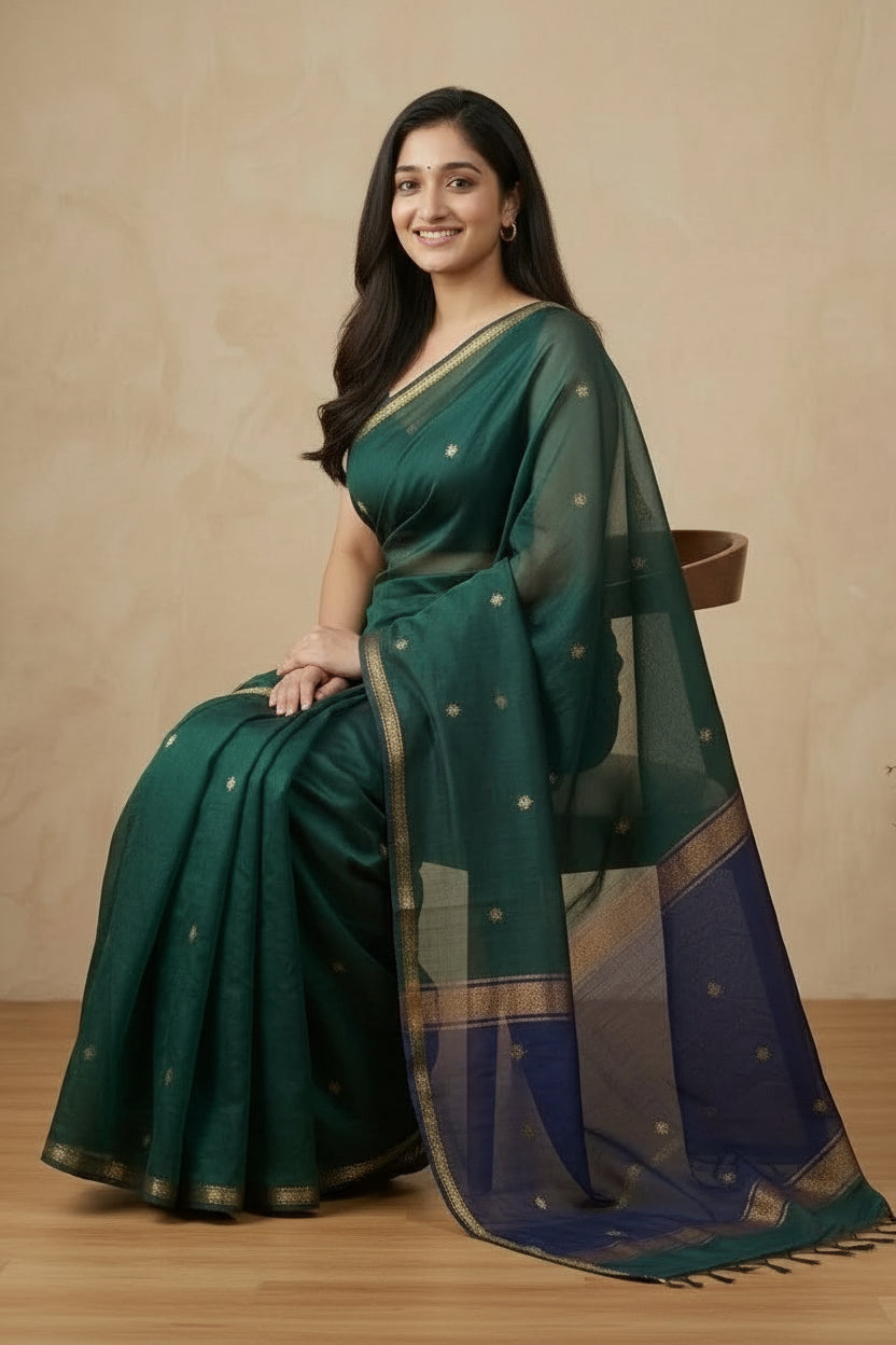 Woman wearing a green and blue saree sitting on a wooden stool against a beige wall.