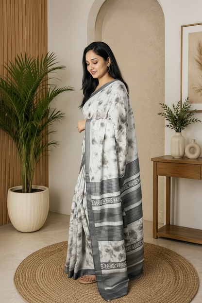 A woman modeling a gray and white tie-dye saree with a patterned border, standing on a round jute rug in a modern, neutral-toned indoor setting.